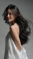 Beauty asian brunette girl with long and shiny wavy hair . Beautiful woman model with curly hairstyle shine and volume in white dress in shiny white background, hair care products and luxury salons.