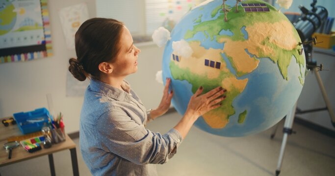 STEM Class: Female School Teacher Educating Science and Renewable Energy Sources Using Model of Planet Earth in Classroom. Miniature 3D Models of Wind Turbines and Solar Panels on Globe. High Angle. - Powered by Adobe
