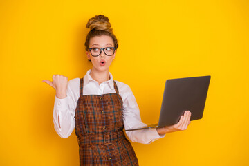 Cheerful young student holding laptop and gesturing, wearing trendy school outfit against bright...