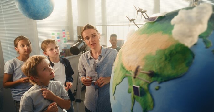 Ecology STEM Class: Elementary School Boy Attaching Miniature Model of Wind Turbine to Globe. Female Teacher Explaining Science and Renewable Energy Sources to Smart Students in Modern Classroom. - Powered by Adobe