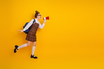 Young female student holding a megaphone and joyfully gesturing, wearing school uniform, standing against a bright yellow background