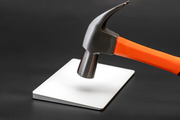 Hammer with orange handle is poised above metal plate on black surface, suggesting impending impact. scene conveys sense of anticipation and focus