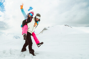 Joyful young couple walk piggyback in deep snow pose looking at camera in snowy mountain,...
