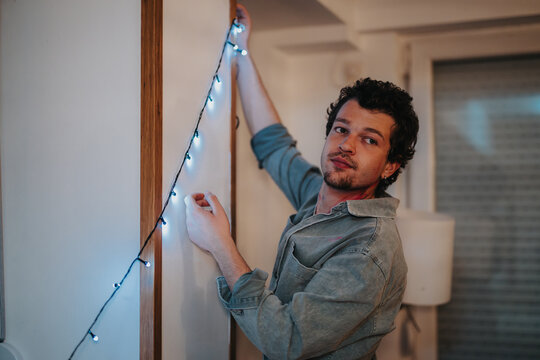 A man in casual attire hangs string lights in a room, preparing for a festive celebration. His focused expression suggests attention to detail, creating a cozy and inviting atmosphere.