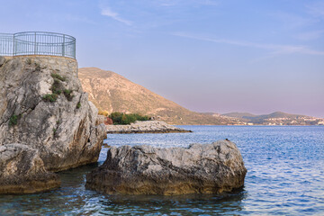 Seaside rock formation with clear blue water and mountains in the background during sunset. Ample...