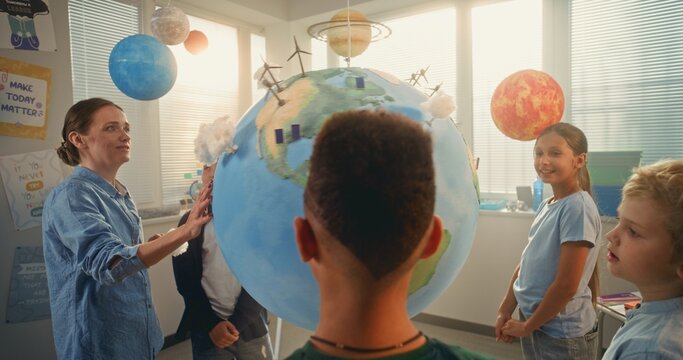Science Lesson: Primary School Children Studying Renewable Energy. Female Teacher Educating Diverse Team of Kids Using Globe with Miniature 3D Models of Wind Turbines and Solar Panels. STEM Education.