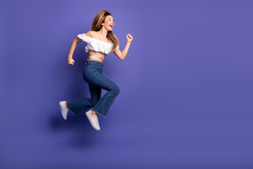Energetic young woman jumping joyfully in casual white top and jeans against a vibrant purple background