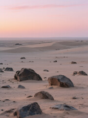 A tranquil desert scene at sunset with soft sand dunes, scattered rocks, and a pastel-colored sky blending into the horizon.