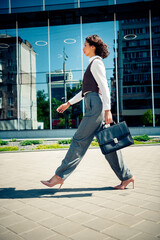 Young businesswoman walking confidently outdoors in urban setting holding briefcase and wearing...