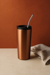 An elegant copper-colored travel mug with a sleek metal straw, resting on a wooden surface with a cloth beside it. The design is minimalistic and modern.