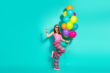 Smiling woman holding colorful balloons on turquoise background, wearing retro hippie fashion, expressing carefree joy