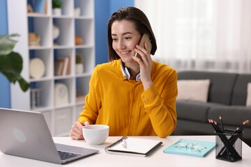 Smiling woman with laptop talking by smartphone at desk in home office