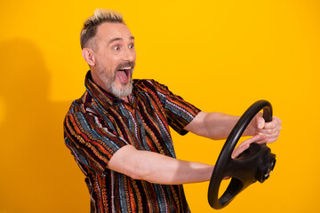 Cheerful middle aged man holding a steering wheel and expressing joy against a yellow background in...