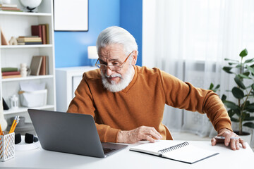 Senior man learning online using laptop at desk indoors. Self-study