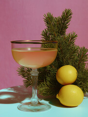 A golden cocktail in a coupe glass with lemons and pine sprigs set against a soft pink background. A minimal festive arrangement blending natural textures and citrus tones.