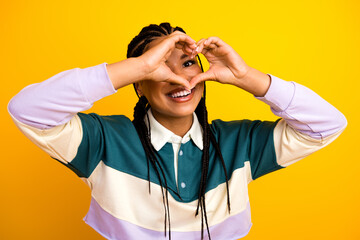 Young woman forming a heart symbol with hands on a yellow background expressing joy, style, and...