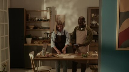 African American senior man with smartphone showing pizza recipe to Caucasian mature wife kneading dough at kitchen table - Powered by Adobe
