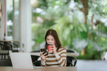 A young woman sits in a bright cafe, focused on her smartphone, with a laptop open in front of her,...