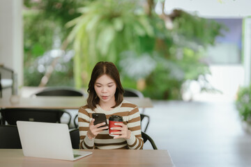 A young woman sits in a bright cafe, focused on her smartphone, with a laptop open in front of her,...
