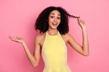Cheerful young woman with curly hair in a stylish yellow dress joyfully holding a strand of hair...