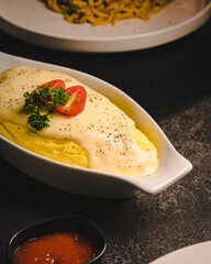 Close-up of a creamy Japanese-style omelet (omurice) topped with rich white sauce, garnished with cherry tomatoes and parsley, served in a white ceramic dish. A small bowl of red sauce is placed nearb