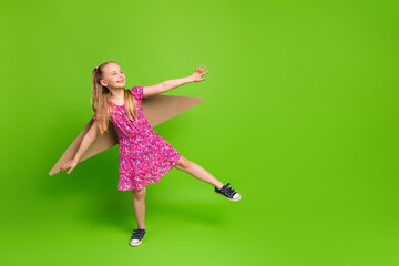 Young girl in playful pose wearing colorful dress with handmade cardboard wings against vibrant green background