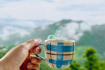 Close-up of a hand holding a steaming hot coffee cup against a scenic mountain backdrop, capturing a peaceful and cozy morning moment in nature.