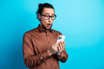 Young man with glasses reacts with surprise holding a smartphone against a turquoise background in...