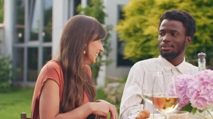 Thoughtful brunette female in colored blouse speaking to attentive African American man in striped shirt. Sitting at table with wine and snacks. Engaging in calm conversation during garden dinner.