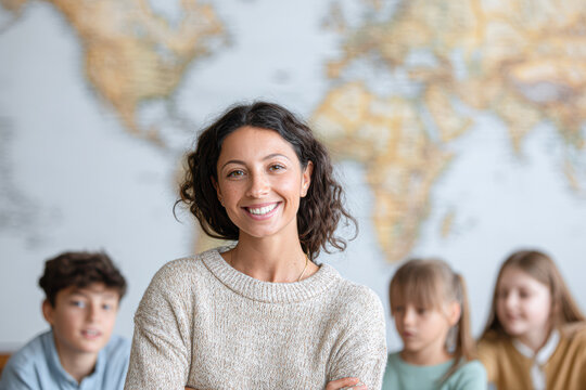 A smiling woman stands confidently in front of a world map, with three children blurred in the background, suggesting a classroom or educational setting.