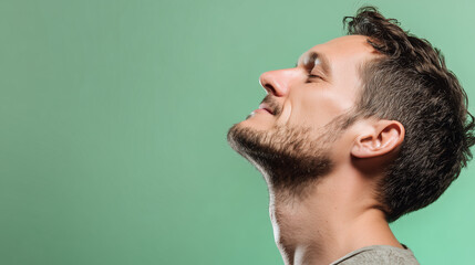 Fototapeta premium Man with beard closing eyes and exhaling deeply with relaxed expression against green background, showing calmness and stress relief in close up side profile portrait