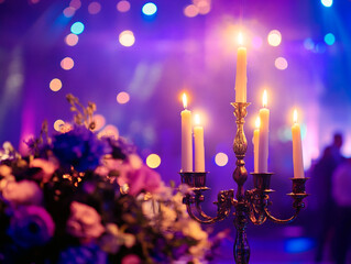 A close-up of a candelabra with lit white candles next to a bouquet of pink and white flowers, all set against a blurry purple background with bokeh lights.