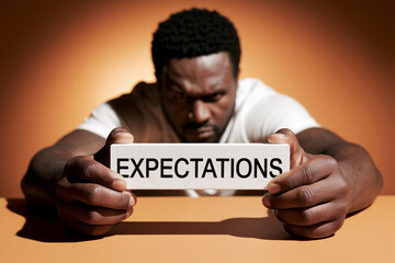 Black man holding block labeled "expectations" in orange studio, symbolizing social pressure and masculine burden