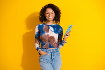 Smiling young woman with smartphone against vibrant yellow background wearing trendy casual outfit with curly hair