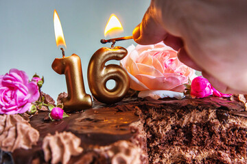 Closeup of a womans hand lighting golden 16 candles on a chocolate birthday cake decorated with...