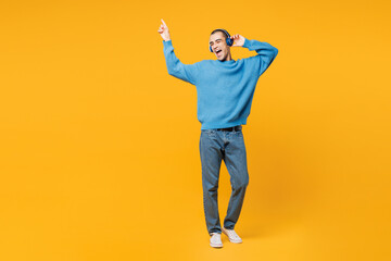 Full body young middle eastern man wearing blue sweatshirt casual clothes listening to music in headphones raising up hands dance isolated on plain yellow orange background studio. Lifestyle concept.