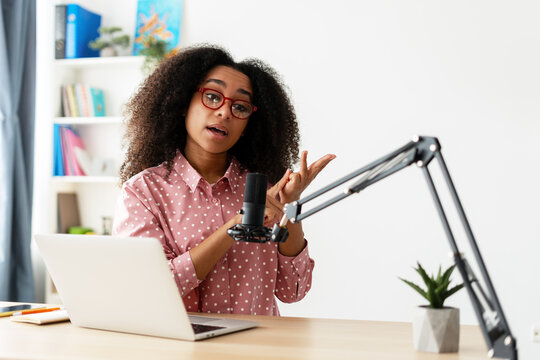 Young curly African American woman recording podcast in home studio using microphone and laptop