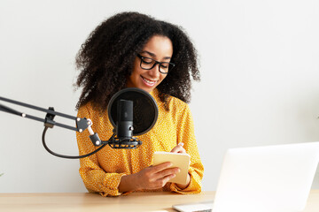 Beautiful African American woman wearing stylish yellow shirt recording podcast and taking notes