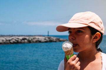 A girl is eating an ice cream cone by the water. The water is blue and the sky is clear
