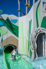 A child is sliding down a water slide. The slide is green and the water is clear. The child is wearing a life jacket