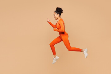 Full body young fitness trainer instructor sporty woman sportsman wear orange clothes spend time in gym jump high run fast look aside isolated on plain beige background. Workout sport fit abs concept.