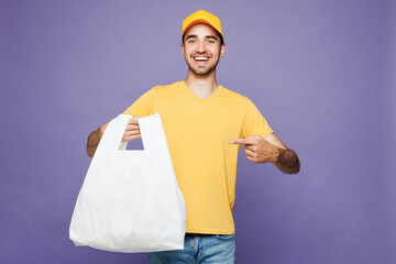 Young delivery guy employee man wears yellow cap t-shirt uniform workwear work as dealer courier hold in hand point finger on plastic bag isolated on plain pastel purple background. Service concept.