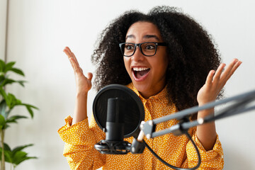 Excited young African American woman wearing eyeglasses recording podcast using microphone