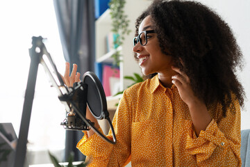Young African American woman wearing stylish eyeglasses recording podcast in home studio