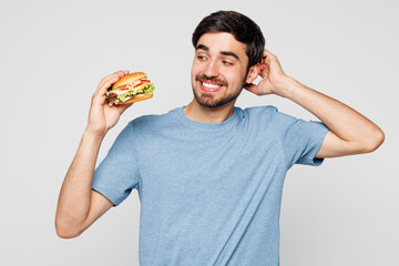 Young tempted minded confused sad man wears blue t-shirt casual clothes hold eat burger scratch...