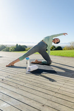 Senior woman performing side-bending on yoga mat on deck overlooking lawn with towel  and  water bot