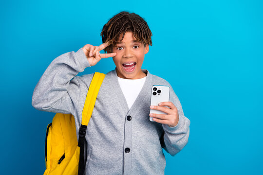 Happy schoolboy posing with a smartphone and yellow backpack on bright blue background showing a cheerful hand gesture - Powered by Adobe