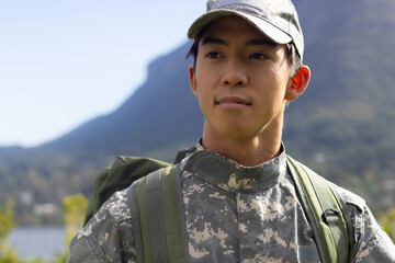 Asian male soldier in camo cap standing by lake near mountains carrying green backpack, copy space