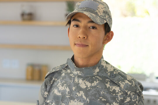 Asian male soldier standing in kitchen by shelves with jars wearing digital camouflage uniform cap