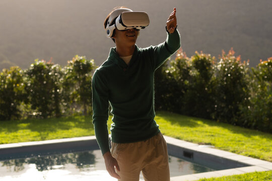 Man wearing VR headset with headphones reaching beside pool on lawn in sunlight, hedge backdrop
